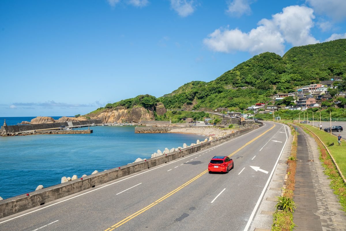 Red car driving along a coastal highway with green hills and blue sea.