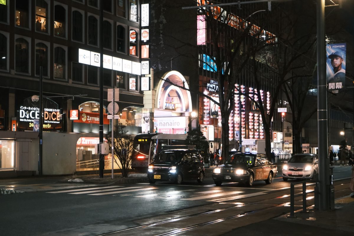 Nighttime city street in Sapporo with glowing storefronts and traffic.