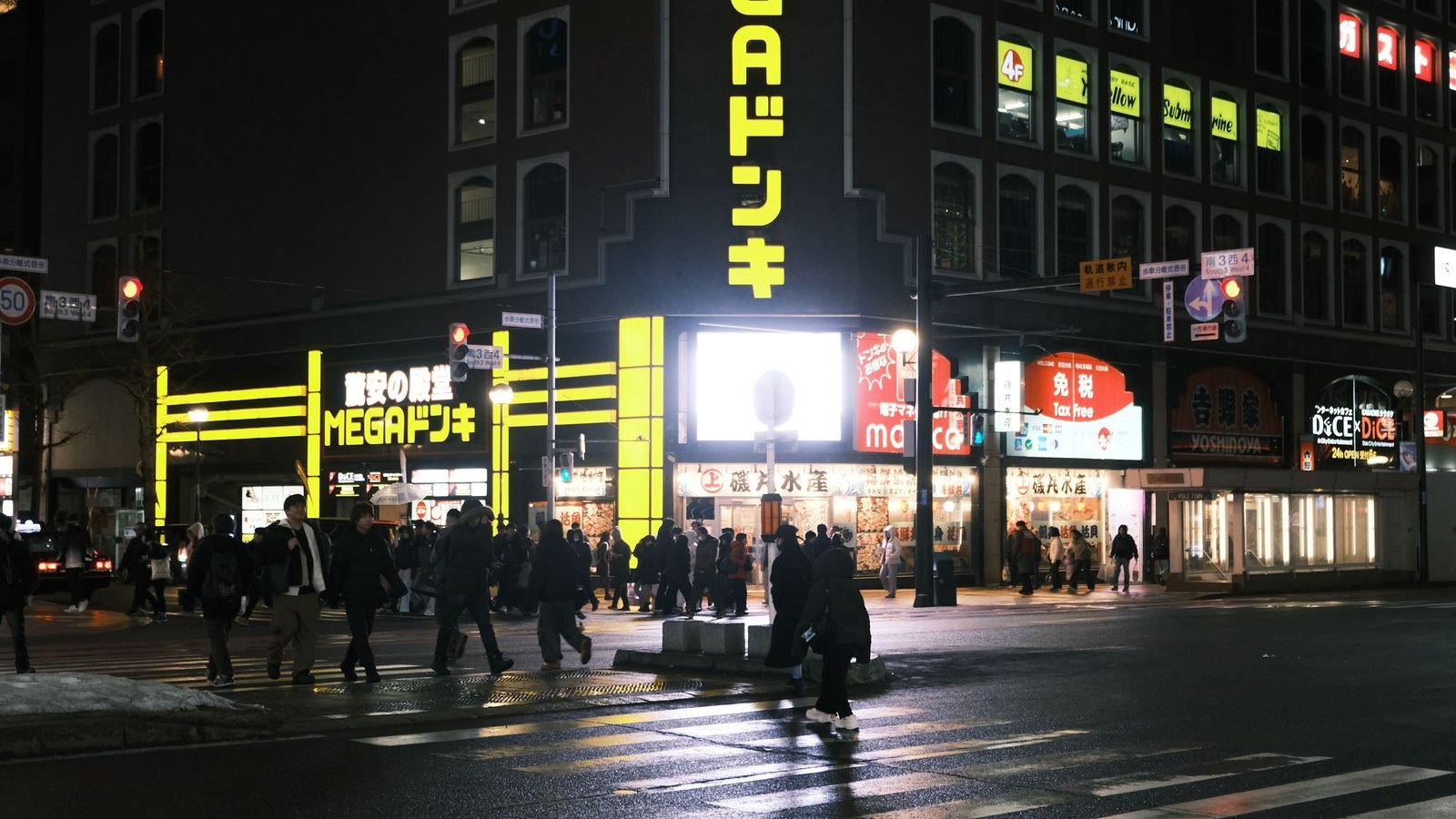 A busy city corner in Sapporo at night with glowing signs and pedestrians crossing.