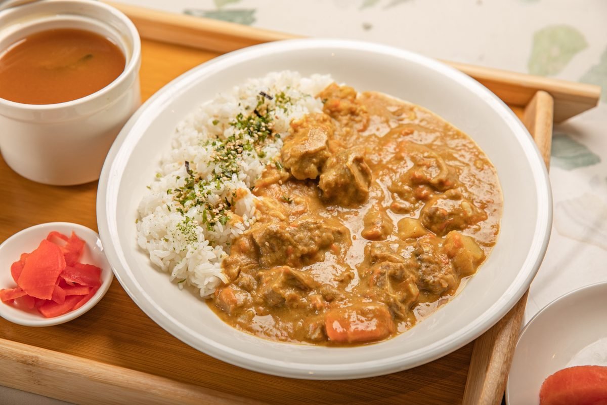 A bowl of Japanese curry with rice, soup, and pickled ginger on a wooden tray.