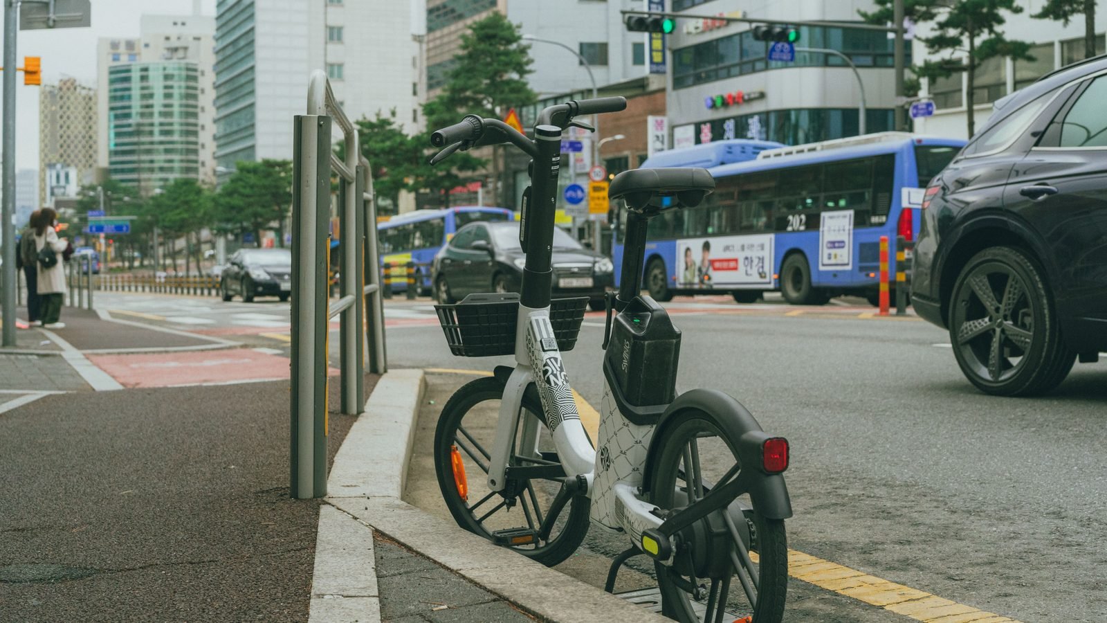 A white and black rental bicycle parked on a Seoul sidewalk with street traffic in the background.