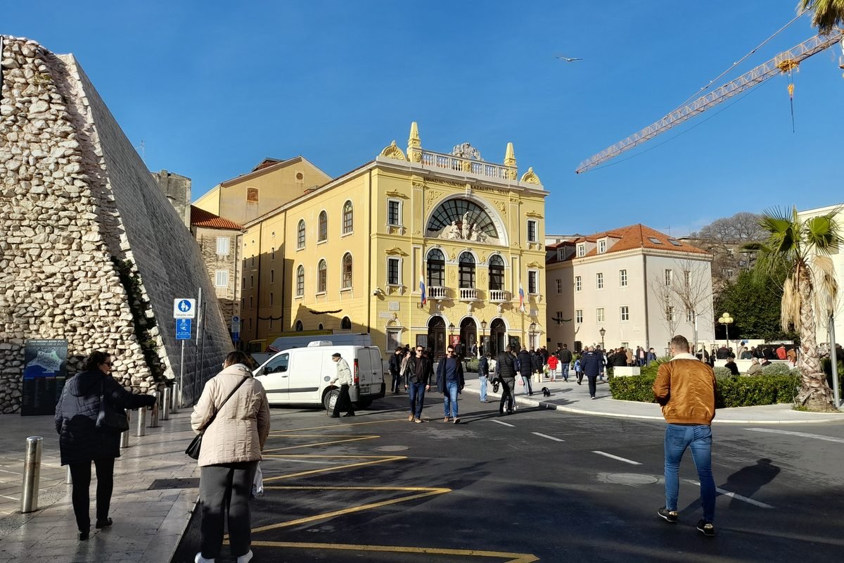 Pedestrians on a European city street near a historic yellow theater building