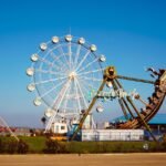An illuminated amusement park Ferris wheel and pirate ship ride near an ocean walkway.