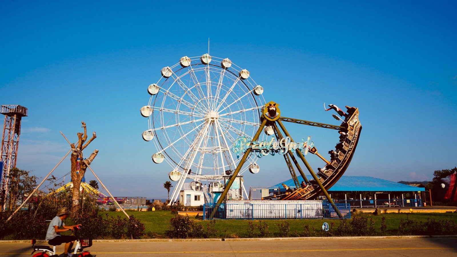 An illuminated amusement park Ferris wheel and pirate ship ride near an ocean walkway.