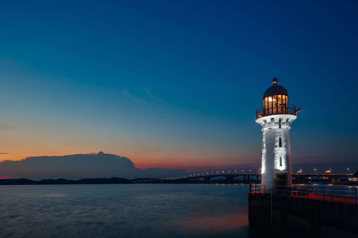 Illuminated lighthouse by a bridge at twilight over calm water.