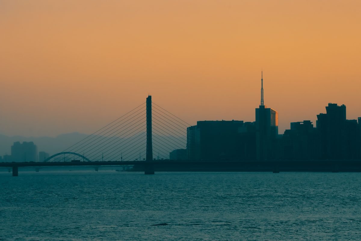 Silhouette of a city skyline and bridge at sunset over water.